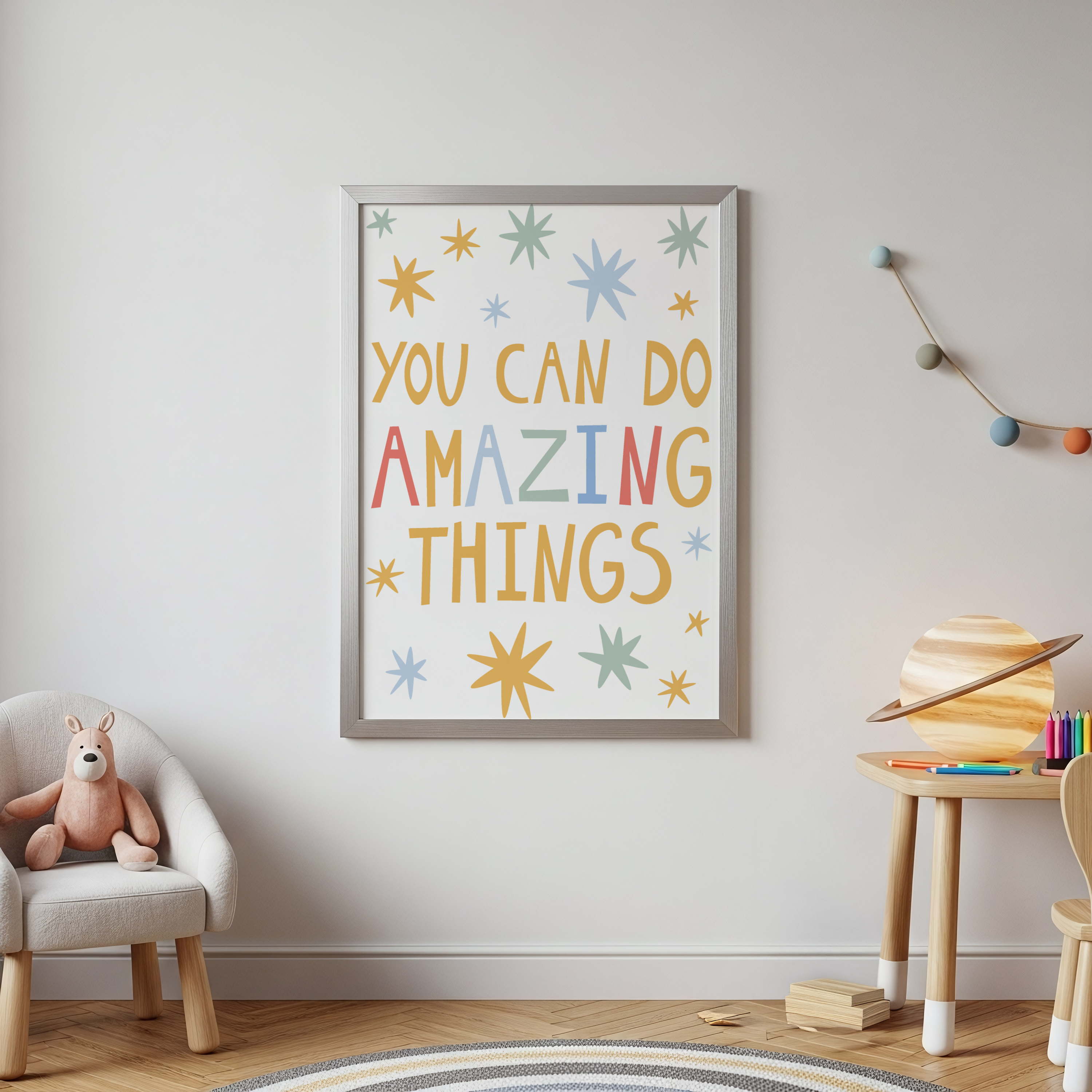 A framed children's wall art print with the text 'YOU CAN DO AMAZING THINGS' in colorful lettering, surrounded by star and burst shapes, displayed in a styled nursery room setting.